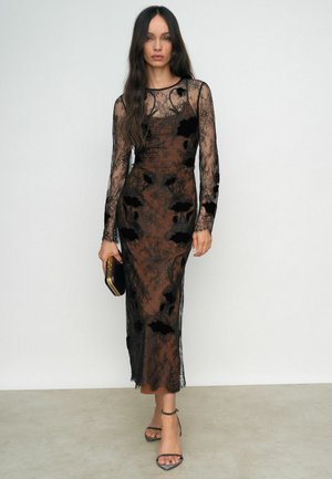 Abito in pizzo nero con maniche lunghe trasparenti, motivi floreali e silhouette aderente. Abbinato a una pochette nera e sandali con cinturino.