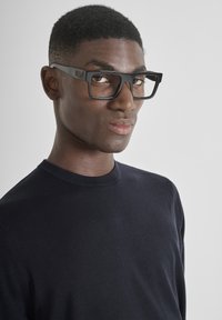 Occhiali neri rettangolari con montatura spessa, indossati da una persona in un maglione blu navy a trama, su uno sfondo grigio chiaro.