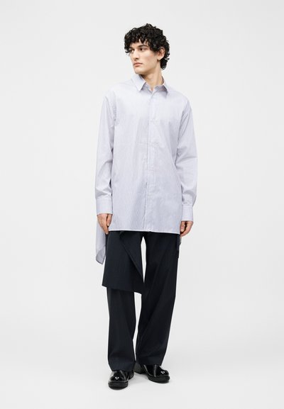 Junger Mann steht, trägt ein hellblau gestreiftes langes Hemd und schwarze asymmetrische Hose mit weitem Bein sowie schwarze Schuhe, blickt zur Seite.