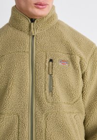 Fleece jacket in olive green, featuring a high collar, zippered front, and chest pocket with logo. Textured, soft fabric with stitched accents.