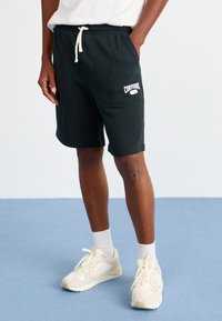 Black cotton shorts with a drawstring waist and side pockets, featuring white "Converse" logo. Model wears light-colored sneakers.