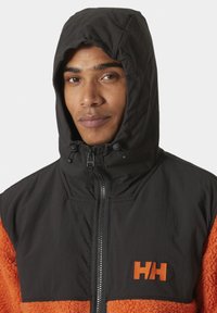 Černá a oranžová bunda s předním zipem, vybavená kapucí, polar fleecovým materiálem na dolní části a vyšívaným logem na hrudi.