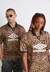 Du grindai jauni suaugusieji, vilkintys atitinkančias rudas leopardų raštus marškinėlius "Umbro"; vienas turi akinius nuo saulės ir trumpus plaukus, kitas - ilgus pintus plaukus ir maišą.