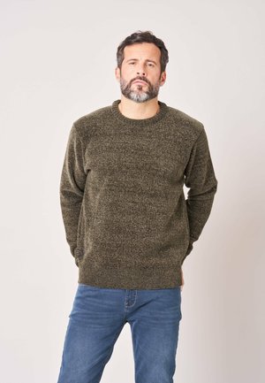 Man met donker haar en baard, draagt een olijfgroen gebreide trui en blauwe spijkerbroek, staand met handen achter de rug, neutrale achtergrond.