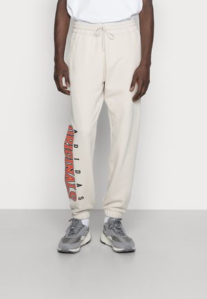 Mężczyzna w białych spodniach dresowych Adidas Originals z czerwonym napisem logo na lewej nogawce i szarych sneakersach z białymi podeszwami.