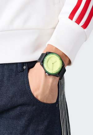 Nadgarstek z czarnym zegarkiem z neonowozieloną tarczą, ręka w kieszeni ciemnoniebieskich jeansów, w białej bluzie z długim rękawem w czerwone paski.