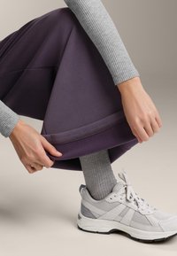 Fialové sportovní kalhoty s širokým střihem se nyní upravují. Také je vidět šedý pletený top s dlouhým rukávem a světle šedé tenisky.