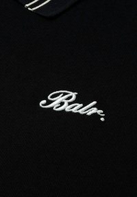 Svart polo skjorta med ribbad textur, som har en vit broderad "Balr."-logga och en kontrasterande rands detaljer på kragen.