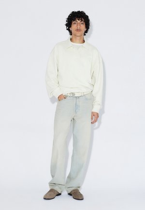 Polo oversize à manches longues couleur crème avec col, associé à un jean droit bleu clair et des chaussures beige à enfiler. Détail de logo subtil.