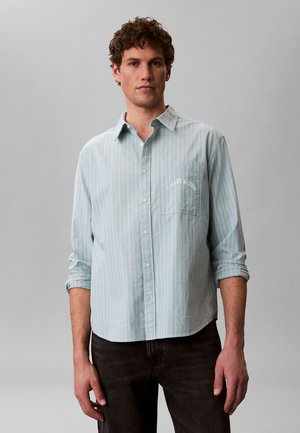 Mann trägt hellblaues, vertikal gestreiftes Hemd mit hochgekrempelten Ärmeln und dunkle Hose vor einfarbigem grauem Hintergrund.