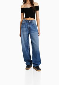 Femme portant un crop top noir dénudé des épaules, un jean bleu taille haute à jambes larges et des baskets noires avec des semelles beiges, se tenant sur un fond blanc.