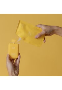 Bottiglia di detergente per mani gialla con tappo a ribalta tenuta in una mano, mentre un sacchetto di ricarica dello stesso giallo viene versato al suo interno.