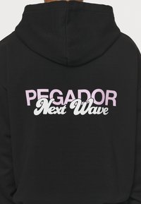 Felpa nera con cappuccio caratterizzata da una grande stampa grafica sul retro con la scritta "PEGADOR Next Wave," in una tipografia grassetto rosa e bianca.