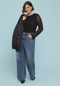 Schwarzes Langarmoberteil mit transparenten Ärmeln, kombiniert mit hochgeschnittenen blauen weiter geschnittenen Jeans und einer schwarzen Bomberjacke, vor einem hellblauen Hintergrund.