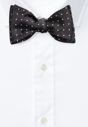 Nœud papillon en soie noire à petits pois blancs sur une chemise blanche avec boutons visibles.
