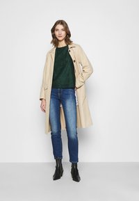 Casaco trench bege por cima de uma blusa verde escura texturizada, combinado com jeans azuis e botas de tornozelo pretas. Apresenta um fecho de botão e corte descontraído.