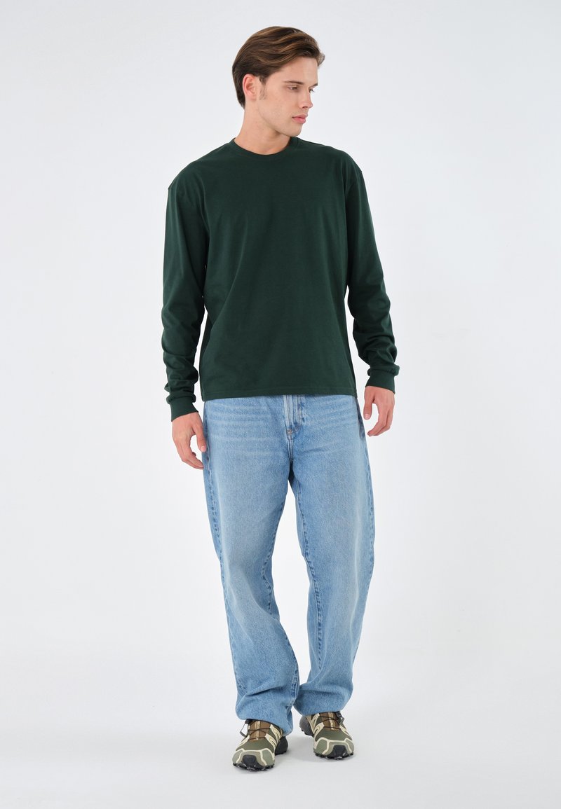 T-shirt à manches longues vert foncé, col rond, associé à un jean bleu clair. Porté avec des baskets à motifs verts et beiges sur un fond neutre.