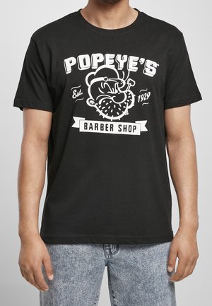 Schwarzes T-Shirt mit weißer Grafik von Popeyes Gesicht, der eine Pfeife raucht, Text "Popeye's Barber Shop Est. 1929", getragen von einer Person in hellblauen Jeans.