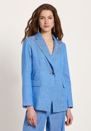 Woman with long brown hair wearing a single-button light blue blazer and matching trousers against a plain background.