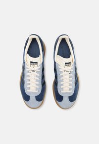 Pár světle a tmavě modrých Adidas tenisek s bílými tkaničkami, gumovými podrážkami a logem Adidas na jazyku, viděno z vrchu na bílém pozadí.