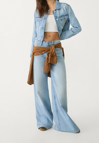 Ljusblå denimjacka i croppad design med två framfickor; kombinerad med vida ljusblå jeans och ett brunt tyg knutet i midjan.