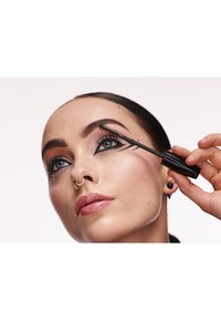 Scovolino di mascara nero applicato alle ciglia. Il soggetto ha sopracciglia definite, labbra nude e un trucco occhi audace con motivi geometrici di eyeliner.