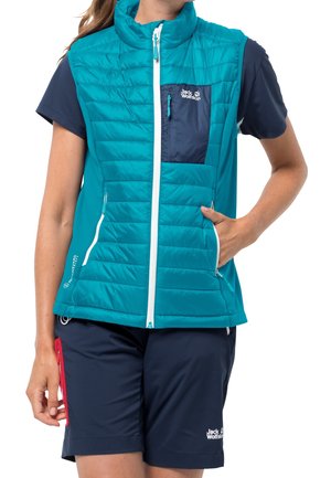 Žena v tyrkysové prošívané vestě bez rukávů s bílým zipem a tmavě modrých šortkách, stojící s jednou rukou v kapse vesty.