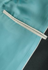 Ein türkisfarbenes, leichtes Gewebe mit glatter Textur zeigt einen kontrastierenden hellgrauen Reißverschluss und Besatz, vor einem dunklen Hintergrund.