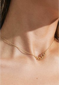 Collana in oro con maglie intrecciate, una delle quali è adornata con piccole pietre trasparenti. La fine catena poggia sulla clavicola, con una finitura in metallo texturizzato.