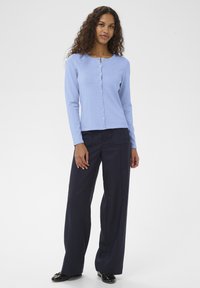 Világoskék bordázott kardigán kerek nyakkivágással és gombos záródással, sötétkék bő szárú nadrággal és fekete cipővel párosítva.