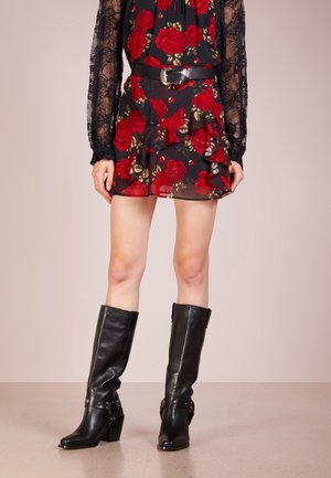 Kvinde iført en sort mininederdel med røde roser, gennemsigtige sorte blonderærmer, et sort bælte og knæhøje sorte læderstøvler.
