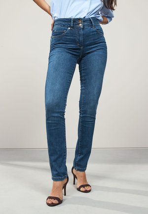 Žena nosiace modré úzke rifle s vysokým pásom a dvojitým zapínaním na gombíky, v kombinácii s čiernymi sandálmi na vysokom opätku s otvorenou špičkou, stojaca na šedom podlahe.