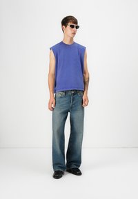 Lila ärmelloses Shirt, distressed blaue Jeans und schwarze Schuhe. Hat einen entspannten Schnitt mit sichtbaren Nähten an den Jeans.