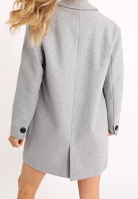 Cappotto grigio in lana, design su misura, monopetto con apertura posteriore, dotato di bottoni neri sulle maniche e colletto a punta.