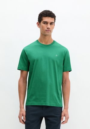 Jeune homme portant un T-shirt vert uni à manches courtes et un pantalon sombre, debout devant un fond blanc, face à l'avant.