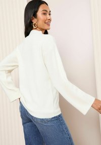 Weißer, weicher Strickpullover mit hohem Kragen und weiten Ärmeln. Kombiniert mit blauen Jeans. Das Model posiert vor einem minimalistischen Hintergrund.