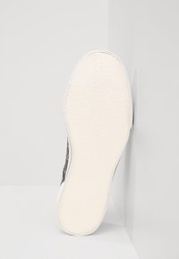 Witte rubberen zool met een textuurpatroon, met daarop een zwart bovenwerk met een geweven ontwerp. Schuine aanblik tegen een lichte achtergrond.