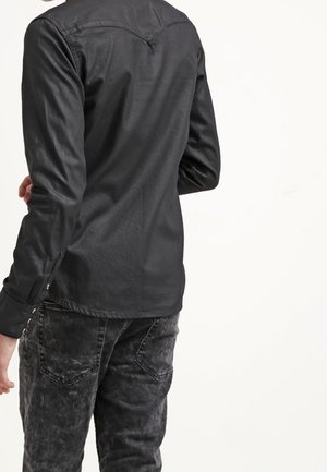 Osoba w dopasowanej czarnej skórzanej koszuli z długim rękawem i czarnych dżinsach z efektem acid wash, odwrócona tyłem do kamery na jednolitym białym tle.