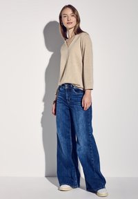Beiger V-Ausschnitt-Pullover mit Dreiviertelärmeln, kombiniert mit blauen weit geschnittenen Jeans. Das Modell trägt weiße Sneakers. Einfache Hintergrund.