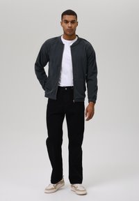 Jachetă bomber din corduroy gri, tricou alb, blugi negri drepti, pantofi sport bej și albi, purtată cu mâinile în buzunare, fundal neutru.