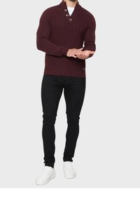 Maglione a collo mezzo in maglia bordeaux con un motivo testurizzato, dotato di tre bottoni. Abbinato a pantaloni neri attillati e sneakers bianche.
