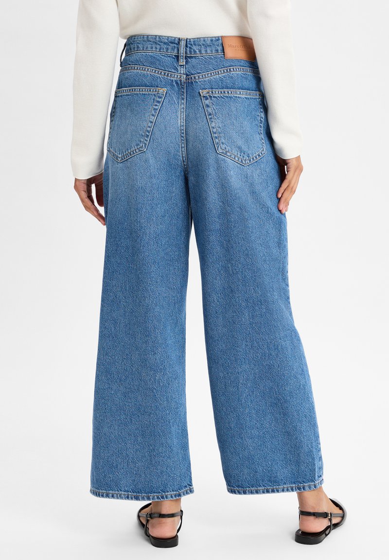 Des jeans en denim bleu à coupe large présentent une taille haute, de grandes poches arrière et une étiquette en cuir à la taille, avec une texture délavée.