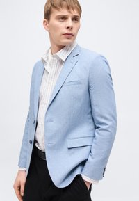 Νεαρός άνδρας με ανοιχτό καστανό μαλλί, φοράει ανοιχτό μπλε σακάκι πάνω από λευκό πουκάμισο με ρίγες και μαύρο παντελόνι, κοιτάζει προς το πλάι με το ένα χέρι στην τσέπη.