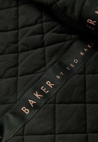Svart kviltat tyg med diagonala stygn; ett satinfärgat band med "BAKER by TED BAKER" präglat i roséguld.