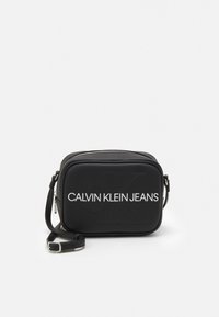 Černá crossbody taška Calvin Klein Jeans s bílým logem a nastavitelným popruhem na bílém pozadí.