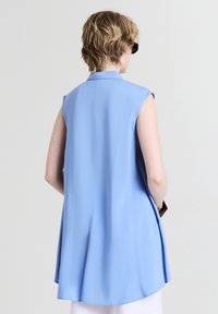 Top blu senza maniche con colletto, silhouette fluida e texture morbida, presenta una schiena dritta e spacchi laterali per facilitare il movimento.