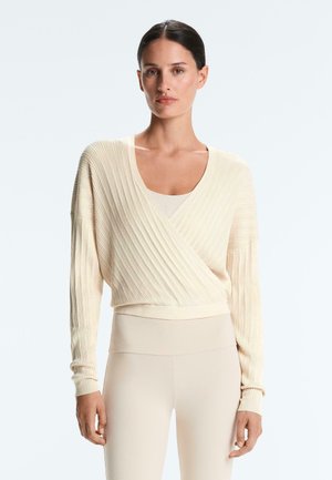 Sötét hajú nő alacsony kontyban, krémszínű bordázott, átkötős pulóvert és magasított derekú bézs leggingset visel, előre néz egy egyszerű háttér előtt.