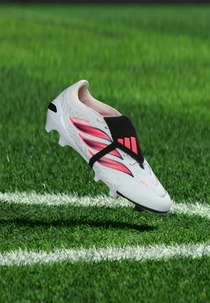 Weißer und rosa Fußballschuh mit schwarzer Abdeckung, der über einem grünen Grasfeld nahe einer weißen Linienmarkierung schwebt.