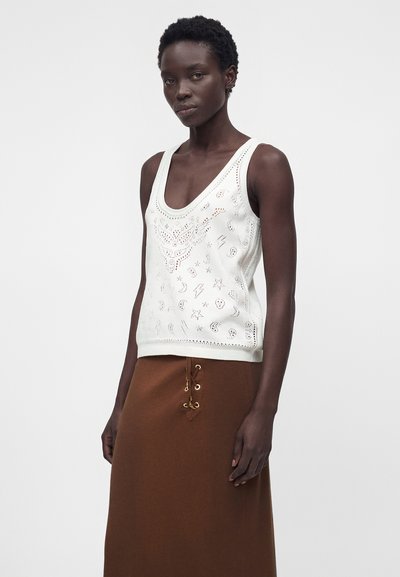 Γυναίκα με λευκό αμάνικο τοπ με κοψίματα και καφέ μίντι φούστα με κορδόνια, στέκεται μπροστά σε απλό φόντο.