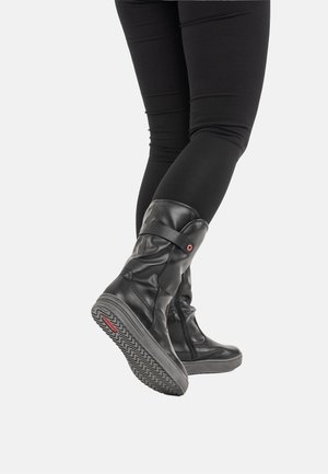 Person wearing black leggings and black mid-calf leather boots with textured sole, one foot slightly raised showing the boot’s bottom.
